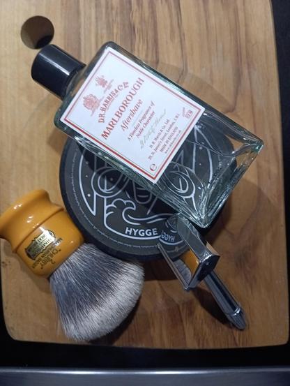 (ChatGPT description)
The image shows a collection of traditional wet shaving items placed on a wooden surface. The items include:
1. A glass bottle of aftershave labeled "DR HARRIS & CO MARLBOROUGH", which appears partially used and features a classic design with a red and white label.
2. A shaving soap container labeled "HYGGE," with a modern, artistic design on the lid.
3. A shaving brush with a yellow handle labeled "Simpson Chubby 2," which has dense bristles.
4. A safety razor with a chrome finish, reflecting light and positioned next to the other shaving accessories.
These items suggest a classic wet shaving setup with a mix of vintage and contemporary products.