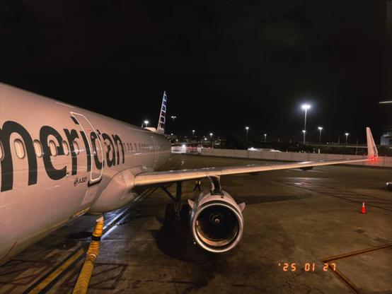 A nighttime scene at an airport terminal, showing the side view of an airplane with the text "merican" visible on the fuselage. The plane is parked on the tarmac with its engine and wing clearly visible. The area is illuminated by several bright lights, and the date "25 01 27" is displayed in the bottom right corner of the image.