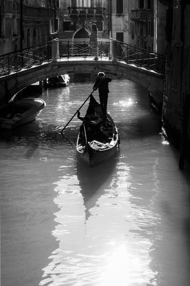 Photography in black and white in which you can see a street scene in Venice (Italy). You can see a traditional boat (Gondola) and also a tourist in a bridge taking a selfie.