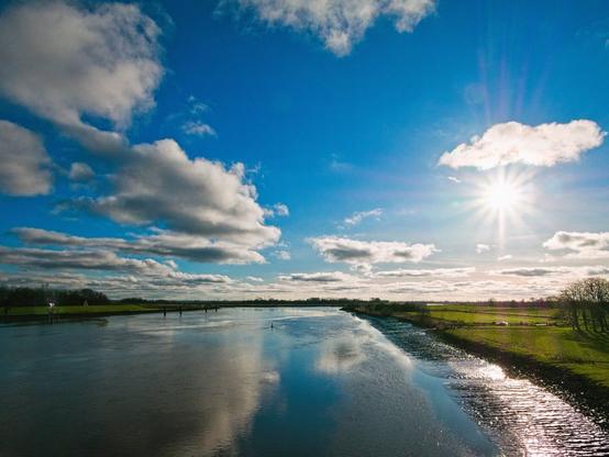 A river lies in front of me. Its waters reflect the blue sky and fluffy, white clouds. The sun is over the banks of the river. There are green meadows and bare trees. It's low tide, so you can see the mud banks on the shore. It is a cold day, but I think everything looks beautiful and clear.
