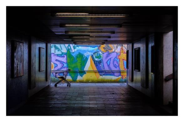 Foto im Querformat. Blick durch eine Unterführung. Rechts geht ein Aufgang hoch, am Ende der Unterführung ist eine Wand, voll mit buntem Graffiti. Davor steht ein leerer Einkaufswagen. Die Unterführung an sich ist dunkel und wir nur minimal von hintereinander gereihten Neonlichtern an der Decke beleuchtet. Die Wand mit Graffiti ist im Tageslicht.