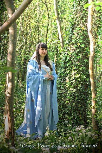 La créatrice d'Atelier Astëria, Claire VINCENT, porte un costume fantasy bleu clair et blanc avec des broderies dorées représentant Dandrane, la soeur du chevalier Perceval des légendes des chevaliers de la table ronde, d'après une illustration de Sarah Bertagna dans le cadre d'une collaboration avec l'illustratrice
