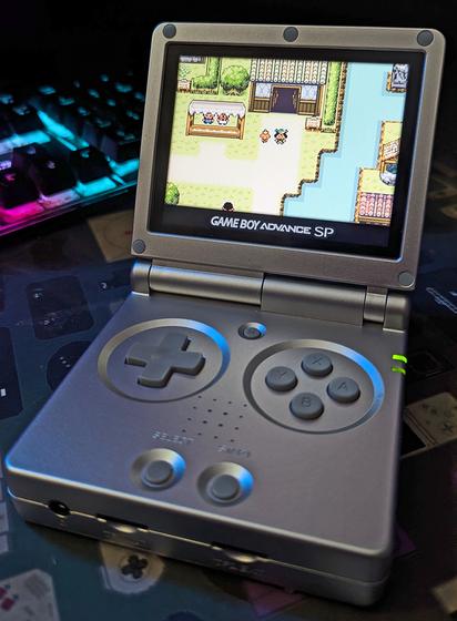 A Gameboy Advance SP looking device playing a ROM hack game. It's Pokémon Emerald but with generation 2 (Gold and Silver) graphics. The player character is standing near a river in a town, with a Torchic by her side.