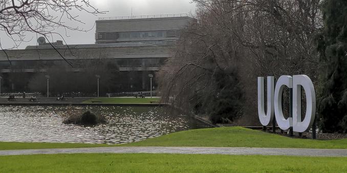 On the right side the UCD letters on campus are in focus with trees behind. On the left you can see a lake with a university building on the opposite side.