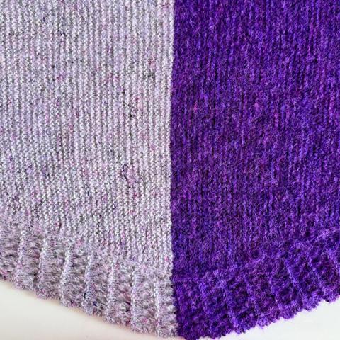 The center of a crescent shaped shawl showing two halves in different colors. On the left is a grey and pink work pink boucle, on the right is purple with neon purple boucle.