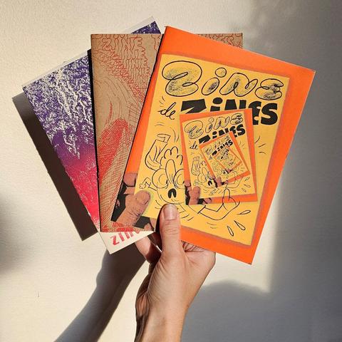 Une main tient 3 zines dans le soleil. On y voit écrit zine de zines .