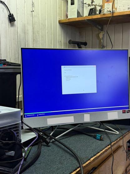 The image shows a computer monitor on a desk in what appears to be a workshop or office setting. The monitor is displaying a Windows installation screen with a blue background. The installation process is underway, and the status window shows a checklist with the following items:

- Copying Windows files
- Getting files ready for installation (37%)
- Installing features
- Installing updates
- Finishing up

There are various cables connected to the monitor, and a device that looks like a projector or a piece of electronic equipment is placed next to it. In the background, there is a wooden shelf with some boxes and tools. The wall behind the monitor is made of vertical wooden panels.