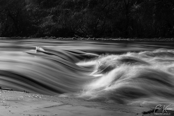 Photo monochrome en noir et blanc captée à une écluse d'un canal en crue avec très peu de dénivelé entre bief supérieur et bief inférieur avec un temps de pose de 0,8 seconde pour lisser les mouvements de l'eau. Le tiers supérieur de la photo est occupé par les arbres de la rive opposée alors que le bas est occupé par le haut de la maçonnerie du sas de l'écluse. Entre les deux, on perçoit la rapidité du courant qui file relativement plat sur le bief supérieur à gauche de la photo et qui est très perturbé après la chute du petit dénivelé restant entre les biefs, générant une vague de retour en rouleau vers l'amont.