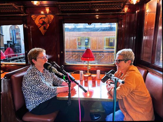 Eva y Sara están sentadas en un antiguo y lujoso vagón cafetería del museo del ferrocarril (Madrid). Luz cálida, maderas nobles, acolchados oscuros. Ellas llevan pelo corto, ropa colorida, gafas. Delante, los micros de grabación. En medio, lamparita de mesa retro.