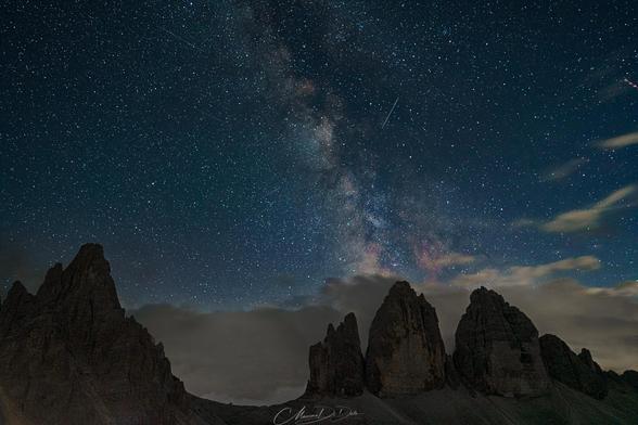 This image shows a breathtaking night sky with the Milky Way galaxy visible in the background. There are rugged mountain peaks silhouetted in the foreground. The sky is filled with numerous stars, and there's a cloud layer beneath the Milky Way, adding to the dramatic composition of the photo.
