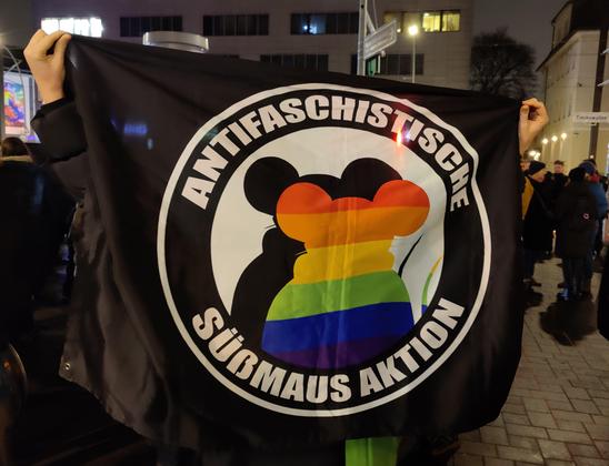 Eine Person breitet hinter ihrem Rücken eine Flagge aus auf der steht "Antifaschistische Saüßmaus Aktion" im Stil der Antifa Flaggen, wo das innere Symbol eine Silhouette einer Maus ist und die normalerweise rot schattierte Maus ist in Regenbogenfarben abgebildet.