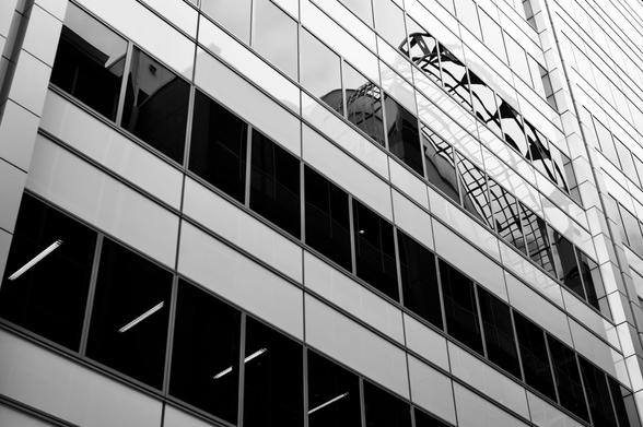 Looking up at the side of a glass and steel building.  A curved steel arch topping the building next door is reflected in the glass windows.