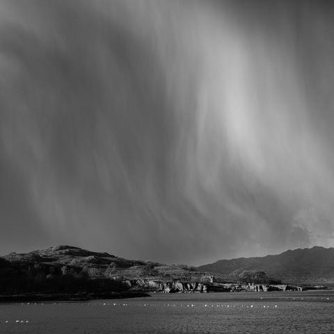 A black and white photo of the top of a rocky peninsula at the left of the frame. It has a small cottage an old fish landing building ruin next to it from back when there were loads of herring in this area. The image is dominated above by a big sky which has sheets of falling hail dropping. Torridon, Wester Ross, Scottish Highlands.

Photo by and copyright of Paul Henni.
