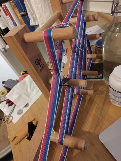 Band-Webrahmen auf einem Tisch, es ist nur noch ca 20 cm unverwobenes Band übrig. Das fertige Band ist in den Fatben der Bi-Flagge (pink lila blau)