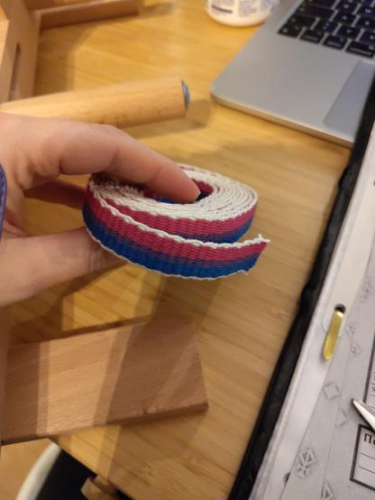 Aufgerolltes hangewobenes Band in den Farben der Bi-Flagge mit weißem rand. Drumherum unordentlicher Schreibtisch.