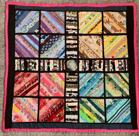 A square string quilt, made from strips of colorful print fabrics, arranged in four diamonds, with a black-and-white striped X through the middle; bordered in black, with a pink binding.