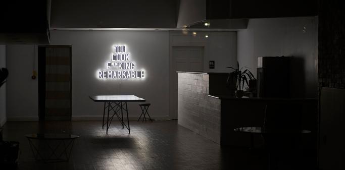 Looking inside of an emptied out business during the height of Covid in 2020,  in the foreground a table eerily appears to levitate in front of a fake neon sign glows telling us that "You look f**king remarkable."