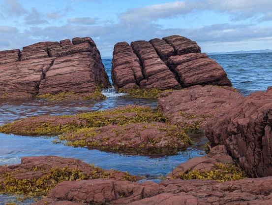 Photo of big squarish chunks of pipestone (i think?) furred with seaweed, sticking up out of peaceful shore water that reflects the very blue sky