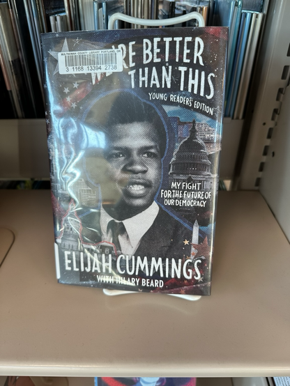 A book entitled “We’re Better Than This: My Fight for the Future of our Democracy—Young Readers’  Edition,  by Elijah Cummings. It is displayed prominently on a library bookshelf.