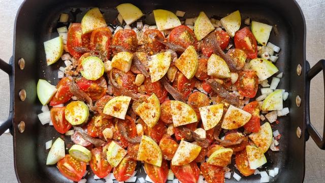 A rectangular, black roasting tray, with pale yellow chunks of zucchini, deep red wedges of tomato, greyish anchovy fillets, garlic cloves and diced onion. Everything has been doused in olive oil, and sprinkled liberally with herbs and spices. The arrangement is almost grid-like.