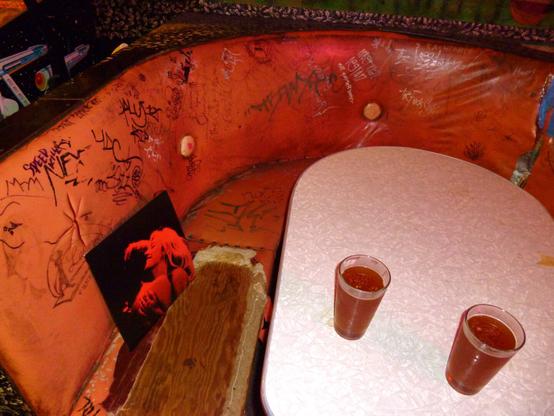 A curved, vinyl booth seat inside the Java Jive is stained, ripped, and covered in black magic market graffiti. A French Exit album by TV Girl is propped up in the seat and two full beers sit on the table.