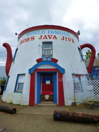 The exterior of the World Famous Java Jive in Tacoma, Washington. The building is shaped like a giant coffee pot, with a handle on one side and a spout on the other, and painted red, white, and blue.