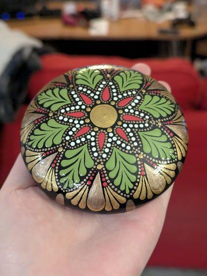 A photo of a dot-painted art stone, with an eight-pointed flower design. The centre dot is gold, the inner petals are a pinkish red and white, and the outer petals are swooshes of pale green. Framing the petals are decadent looking teardrop shapes in gold and bronze. The whole flower design is outlined with bronze dots.