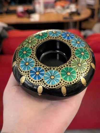 A photo of a round dot-painted tealight candle holder. Circular elements with radial stripes of blue, turquoise, green, and yellow-green encircle the inner well that holds the tealight. The circular elements are surrounded with gold and black dots that look a little like some kind of lace. The hole thing is painted black beneath the design.