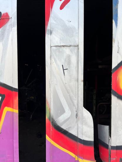 A large industrial garage-type door of aluminum and glass has been spray painted with a graffiti tag so ambitious it covers everything. The tag is silver, red, orange, purple. You can only tell where there’s glass underneath because some panes have broken, leaving broad vertical strips of darkness to interrupt the colour. This is a close-up, two strips of broken window against the paint in between.