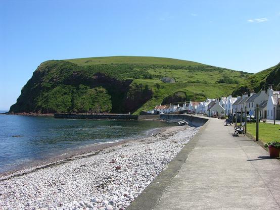 The amazing village of Pennan. The image shows a view along the sea wall with a concrete path on top of it. There's a stony beach to the left descending to the sea, which looks blue under a clear blue sky. To the right is a grassy strip and posts for hanging clothes, then a road and the white cottages and houses of the village. The land rises steeply behind the village and there's a large headland in the background.