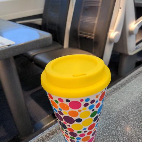 A reusable Coffee cup with an empty seat in the train in the Background.