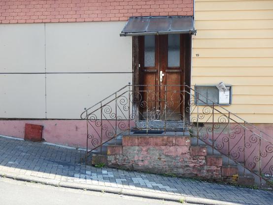 eine alte holztür, eingerahmt von verschiedenen hauswand-verkleidungen, davor ein kleiner beidseitiger treppenaufgang mit schmiedeeisernem geländer