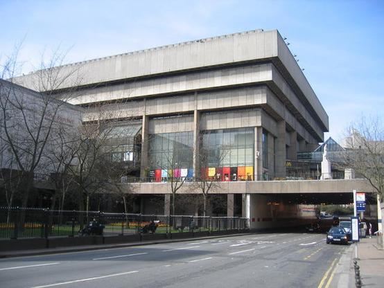 A concrete rectangular building with upper floors jettied outwards.  Sometimes called an inverted ziggurat. The lowest floor has tall glass windows. A decorative coloured band brightens the bleak plain concrete
