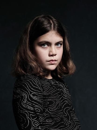 Porträt eines jungen Mädchens mit schulterlangen Haaren, das einen schwarzen Pullover mit silbernem Muster trägt und mit ernster Miene in die Kamera schaut, vor einem dunklen Hintergrund mit weicher Studiobeleuchtung.