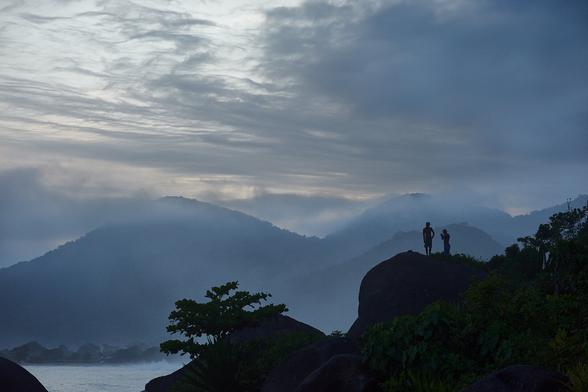 Hills are seen in front of the sea, with two boys / men standing on top of one of them. As the light is behind them they appear dark like shadows. Behind the sea are more hills visible in between clouds. The sun has already set and the rest of the light is rather dim.