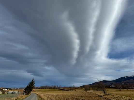 The image shows a dramatic sky with thick, elongated cloud formations stretching across the scene. The landscape includes an open field with a road, a few trees, and hills in the background. The clouds appear to form bands or streaks, creating a striking visual effect.