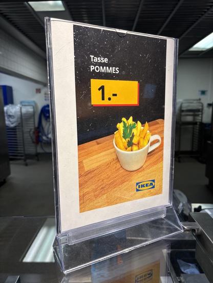 Schild auf einem Tresen.
Abgebildet ist eine Tasse in der Pommes stecken.
Darüber steht: Tasse Pommes 1,-
Darunter steht in der Ecke das Logo von Ikea.
Im Hintergrund ist eine Großküche zu erahnen.