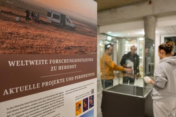 Links im Vordergrund ist eine Ausstellungstafel mit der Überschrift "Weltweite Forschungsinitiativen zu Herodot. Aktuelle Projekte und Perspektiven" zu sehen. Rechts im Hintergrund stehen mehrere Personen um eine Vitrine.