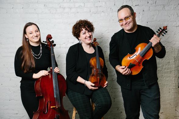 photo of three musicians holding their instruments -- a cello, a violin, and a fadolín (an extended range fiddle). They're wearing mostly black clothes and smiling