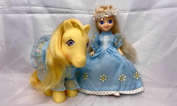 A brightly colored yellow pony and a doll with long blonde hair are positioned side by side. The pony has a flowing mane and a friendly expression, while the doll wears a blue dress adorned with white flowers and lace, complemented by a floral crown.