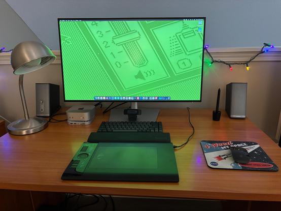 A 32” Dell U-series monitor, two old Bose Companion speakers, an M4 Mac Mini, RayCue base, Satechi keyboard, and old Wacom Intuos4 on a desktop.  The wallpaper looks like old Finder elements.
