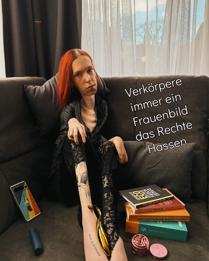 Ich sitze in der Ecke meiner Couch und schaue in die Kamera. Meine schwarze Bluse mit silbernen Details ist halb geöffnet, mein Spitzenrock wird langsam von mir nach oben gezogen. Beides gibt einen Blick auf meine Tattoos frei. zwischen meinen Füßen liegt ein Glasdildo. Links im Bild neben mir liegt ein Vaporizer und mein Handy, von dem die Rückseite zu sehen ist. Die Rückseite zeigt eine Frau vor einem Regenbogenhintergrund die sich ihr T-Shirt über den Kopf zieht und dabei den Mittelfinger ihrer rechten Hand zeigt, indem sie den Arm nach oben hält. Rechts von mir liegen Bücher über Faschismus, trans* Sein, Disability Studies und Rassismus. Außerdem ein Grinder mit einem Stück Kräuter darauf. Neben mir befindet sich noch folgender Text: Verkörpere immer ein Frauenbild das Rechte Hassen.