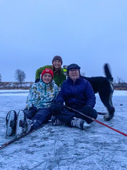 An old man poses for the camera with his two adult daughters and their dog, a large poodle mix. They’re on a frozen lake, and the humans wear winter clothes and ice skates, and hold hockey sticks.