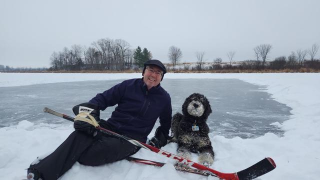 An old man poses with a hockey stick and a large poodle mix, on a frozen lake with a snow-cleared ice rink in the background. The sky is grey.