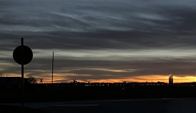 Das Bild zeigt einen Sonnenaufgang über einer leicht industrialisierten Landschaft. Der Himmel ist mit vielen Schichten von Wolken bedeckt, die hauptsächlich dunkelgrau sind, mit einem Streifen orangefarbener und goldener Farben am Horizont, wo die Sonne aufgeht. Im Vordergrund befinden sich die silhouettierten Formen eines runden Verkehrsschilds an einem Pfosten und ein paar schlanke Pfosten. Im Hintergrund sind die silhouettierten Formen von Gebäuden und möglicherweise Industrieanlagen zu sehen, die im nachtbläulichen Licht des Sonnenaufgangs schwach beleuchtet sind. Ein einzelnes, etwas höheres Gebäude ist rechts vom Bild zu erkennen. Die allgemeine Stimmung des Bildes ist düster, aber auch friedlich und atmosphärisch, da die warmen Farben des Sonnenaufgangs einen Kontrast zu den dunklen Silhouetten bilden.

Bereitgestellt von @altbot, generiert mit Gemini