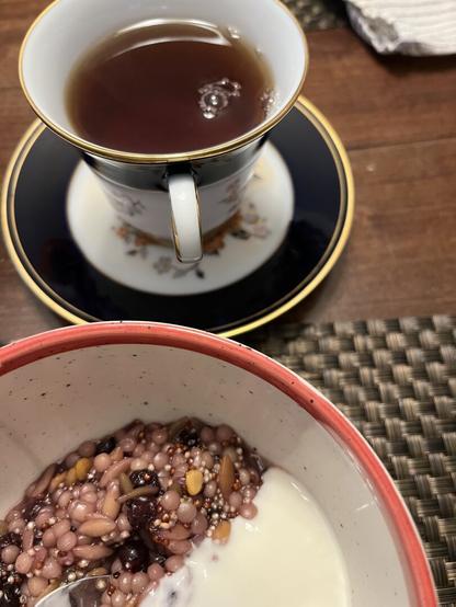A cup of tea sits on a saucer beside a porridge bowl of blueberry and apple cider stained mixed grains with dollop of yogurt
