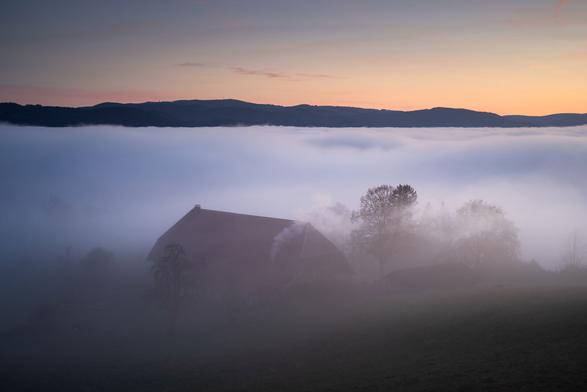 Landschaft in der Abenddämmerung bei Inversionwetterlage. Standort ist genau an der Nebelobergrenze. Unter dem Nebel, gerade noch erkennbar, ein Schwarzwälder Bauernhaus und einige Bäume. Über dem Nebel herrscht klare Sicht auf eine Hügelkette in der Ferne, zu der auch der Feldberg gehört.