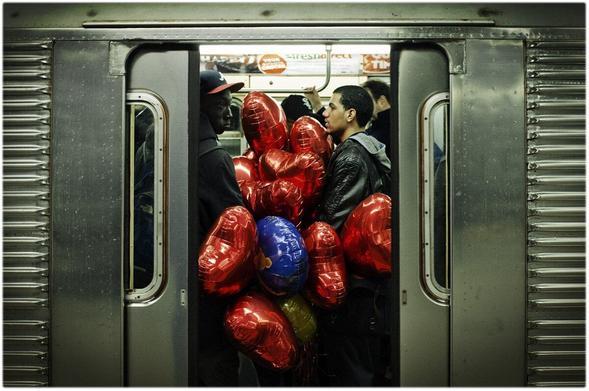 A photograph of a New York City subway car, with the doors closing on a bunch of Valentine's Day balloons. By a bunch, I mean 12 to 15 balloons, in an already crowded car. The gentleman next to the guy carrying in all those balloons is completely unaffected, because this is New York City life. You make room for one more. You deal with less space. And you mind your business.