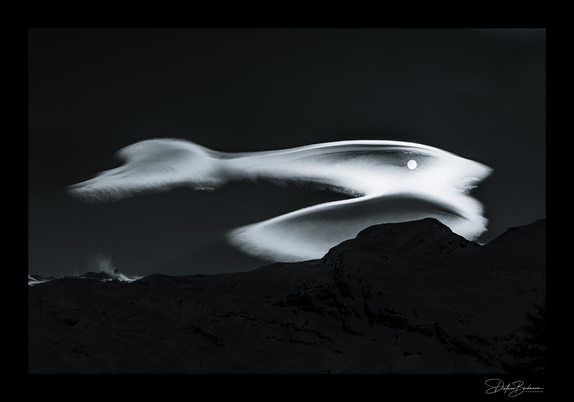 Ein atmosphärisches Schwarzweißfoto einer beeindruckenden Lentikulariswolke über einer dunklen Bergsilhouette. Die Wolke erscheint wie eine elegant geschwungene, weiße Form am dunklen Nachthimmel, die an einen fliegenden Vogel oder eine Welle erinnert. Der helle Mond ist als kleiner weißer Punkt in der oberen Wolkenformation zu erkennen. Die Bergkette im Vordergrund ist als schwarze Silhouette dargestellt, was einen starken Kontrast zur leuchtend weißen Wolke bildet. Die dramatische Komposition und die Lichtverhältnisse verleihen dem Bild eine fast surreale, künstlerische Qualität. In der unteren rechten Ecke befindet sich eine Fotografensignatur.