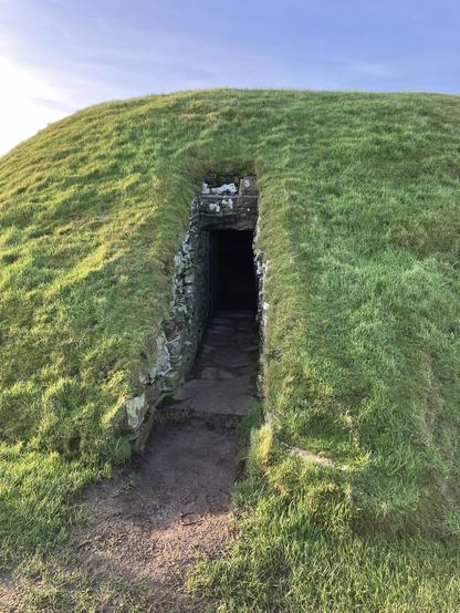 A photo showing the dark, narrow entrance to a tomb that looks like a perfect green hill.